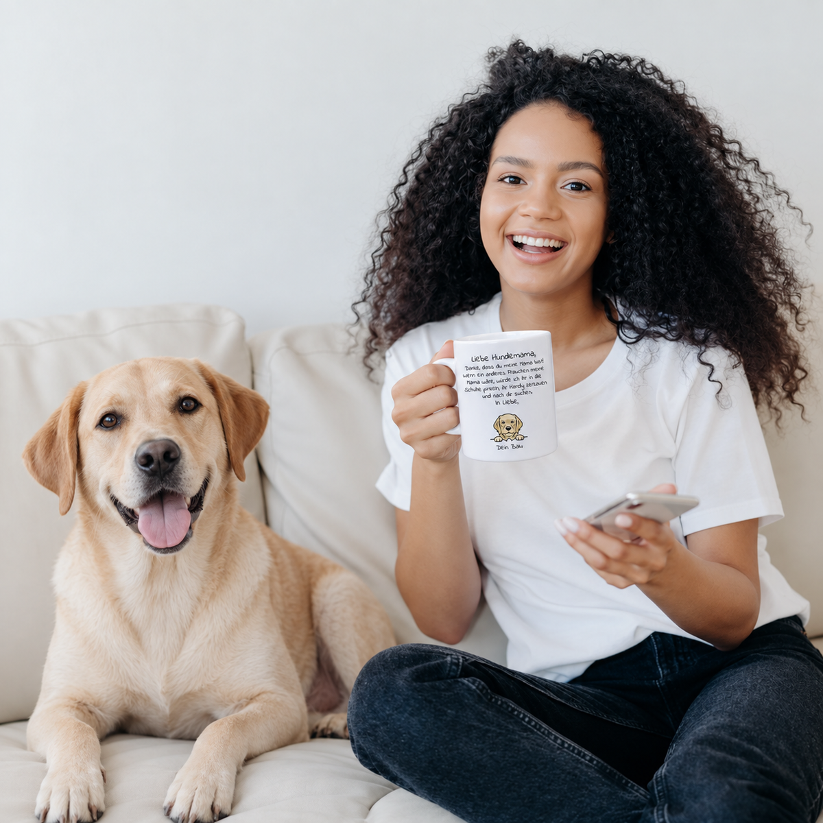 Personne aux cheveux bruns bouclés tenant un mug blanc à motif de chien, à côté d'un labrador sur le canapé.