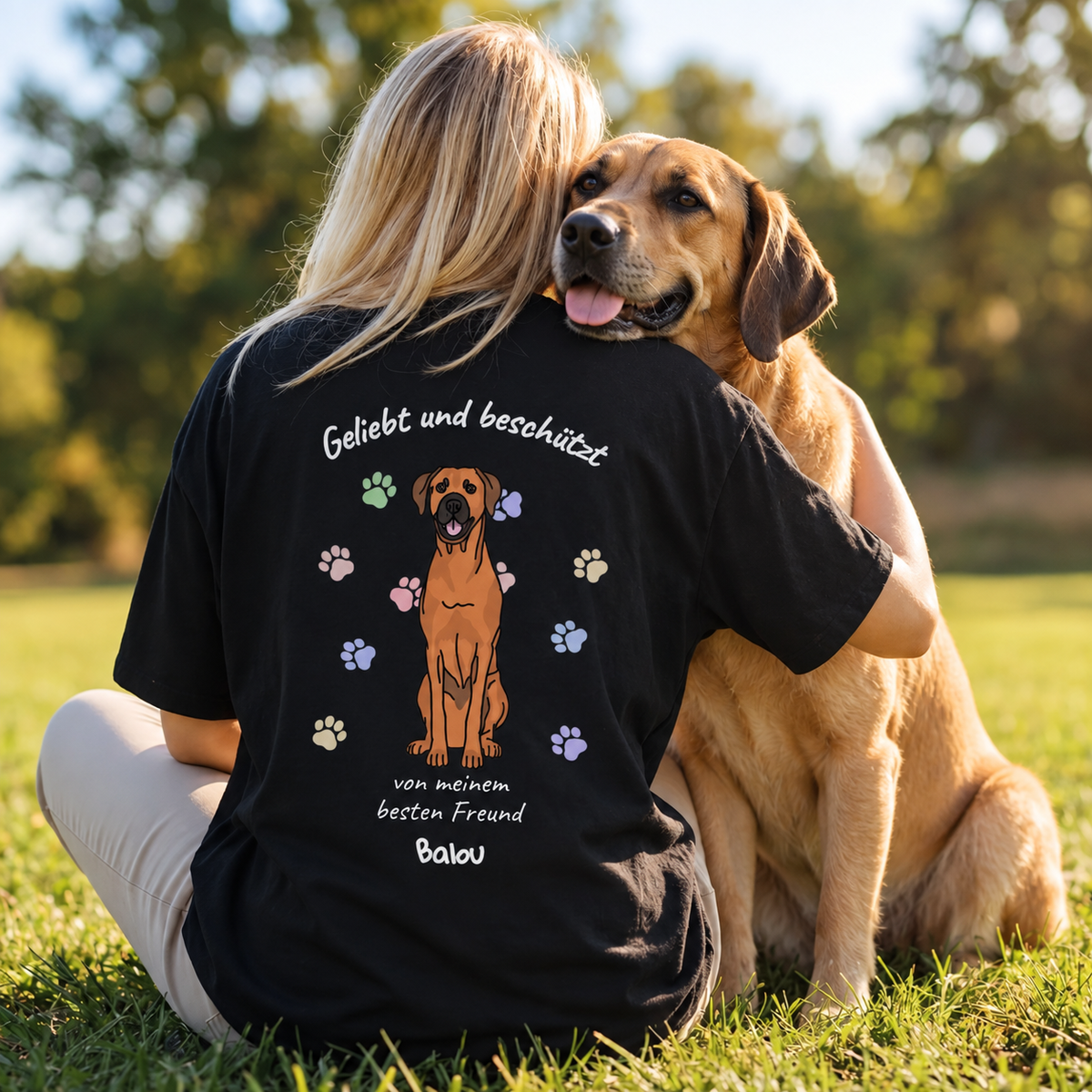 T-shirt noir avec l'inscription "Geliebt und beschützt", un chien et des empreintes colorées.