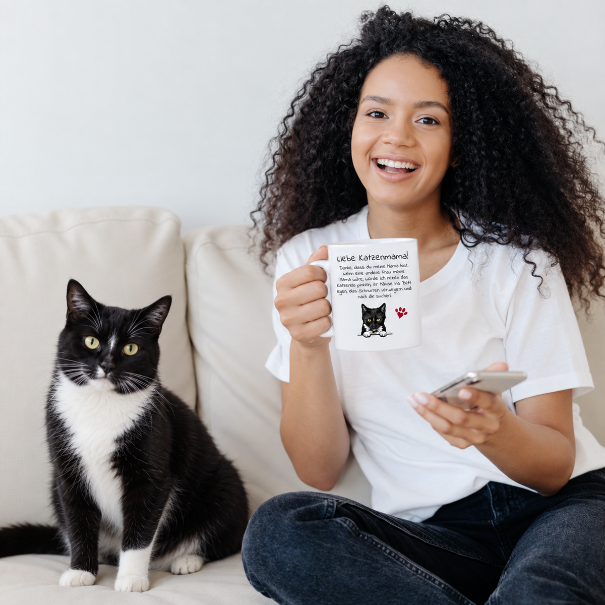 Jeune femme aux cheveux bouclés tenant un mug personnalisé à motif de chat, chat noir assis à côté sur le canapé.