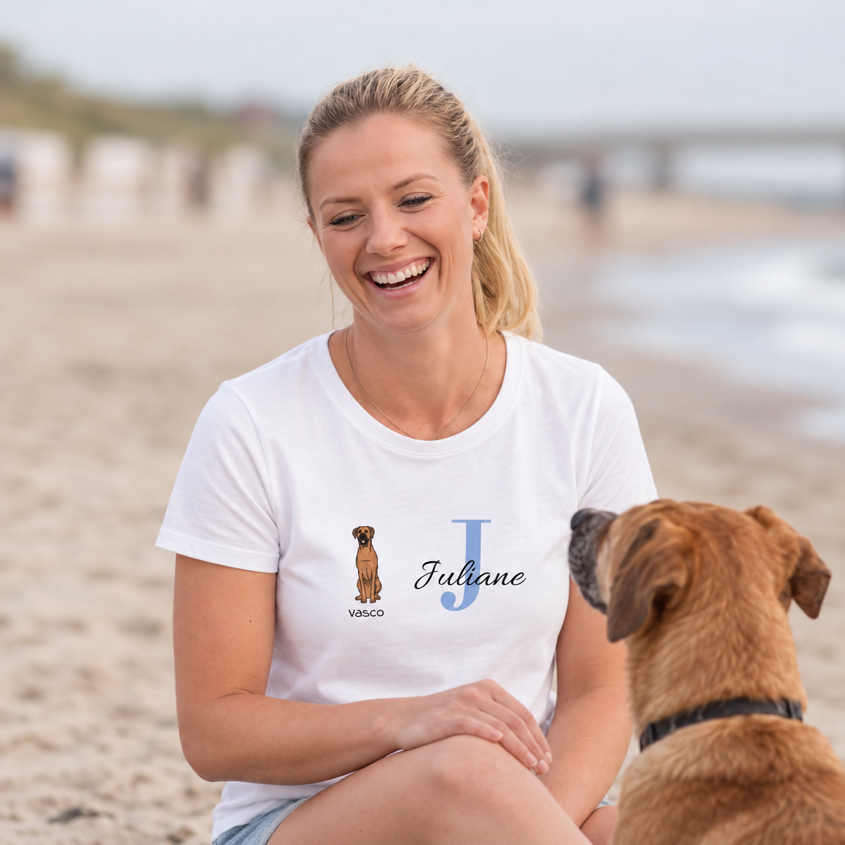 Wit T-shirt met hondenafbeelding en de naam Juliane in blauwe letters. Vrouw zit op het strand.