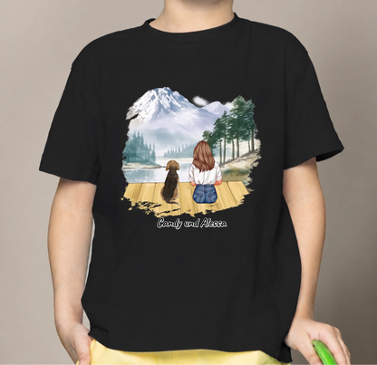 Zwart kinder-T-shirt met afbeelding van een meisje en hond bij een meer met bergen op de achtergrond.
