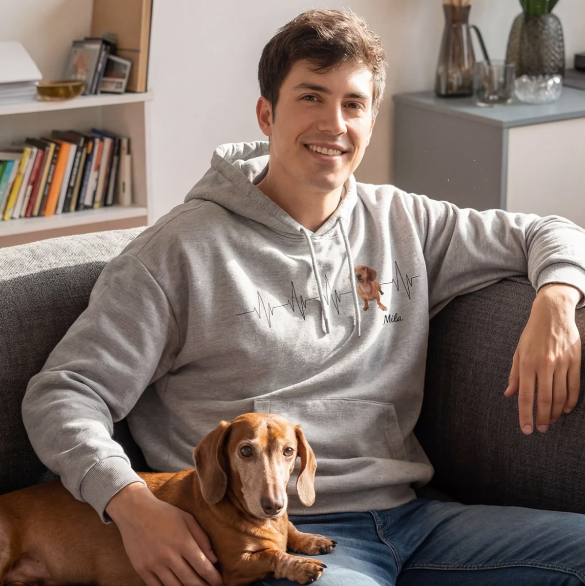 Sweat à capuche gris avec motif battement de cœur et photo de chien, personne assise sur un canapé avec un teckel dans les bras.