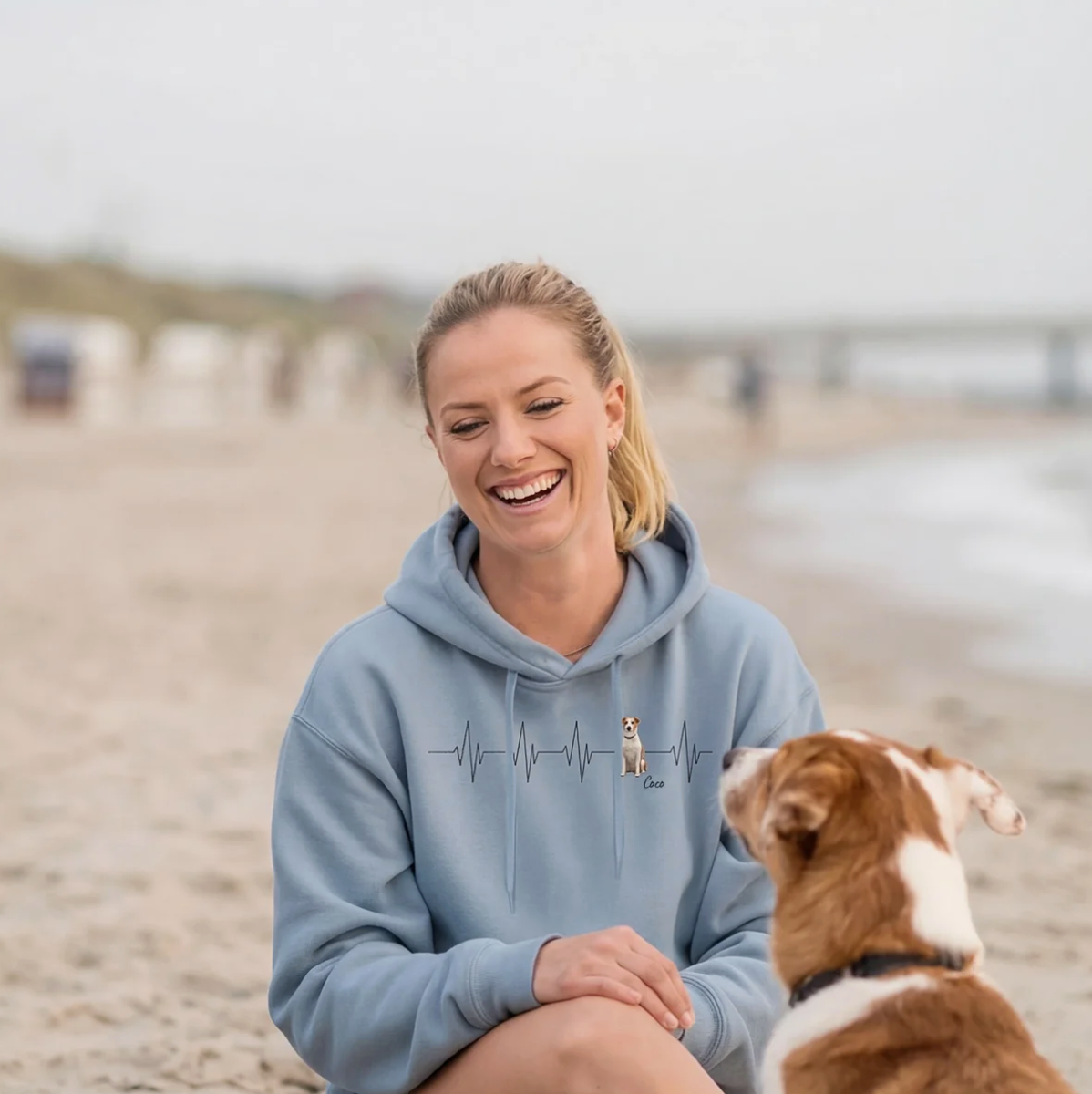 Sweat à capuche bleu personnalisé avec motif battement de cœur et chien, femme souriante assise sur la plage.