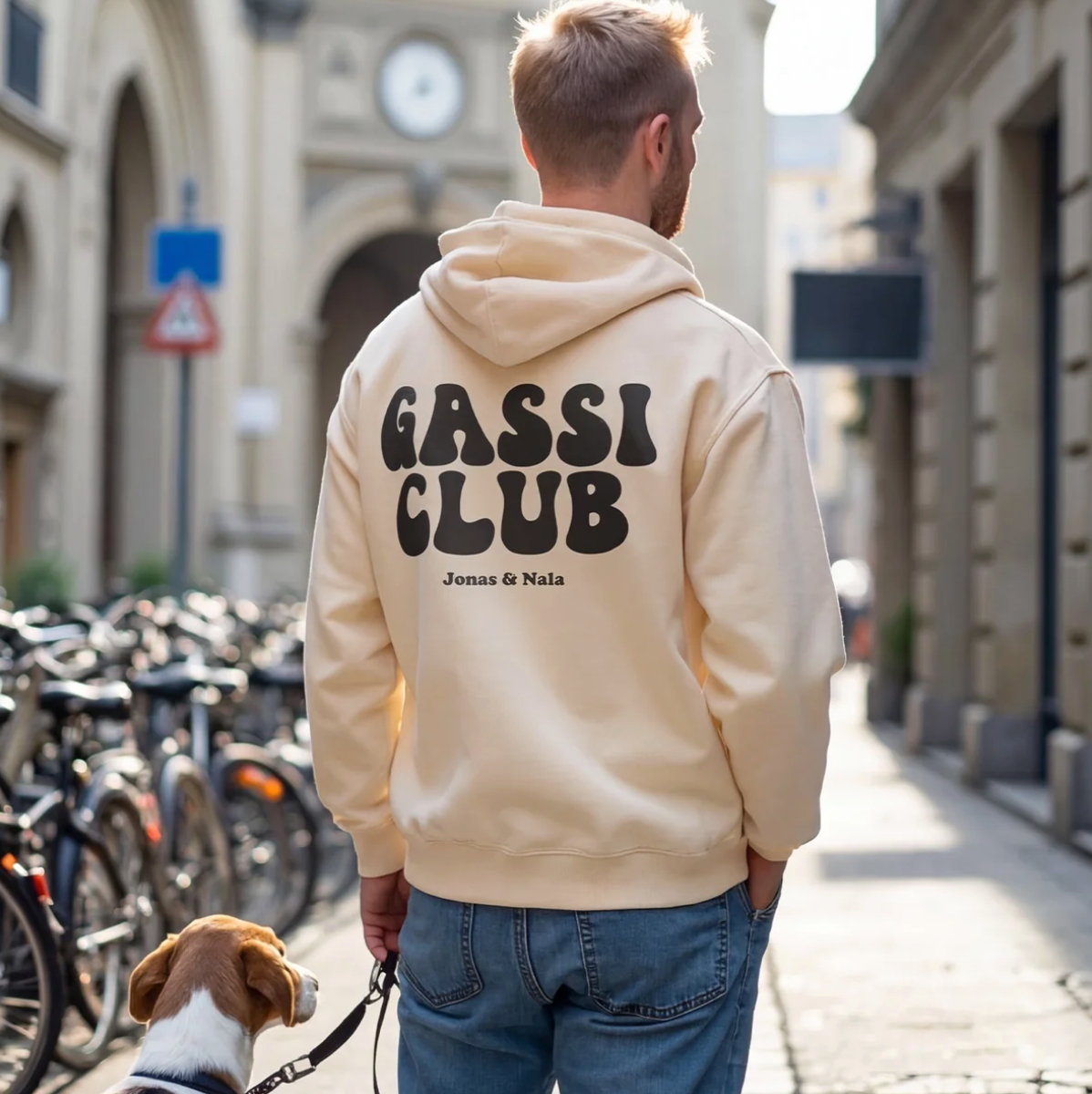 Persoon in een beige hoodie met "Gassi Club" op de rug, staat met een hond in een stadsstraat.