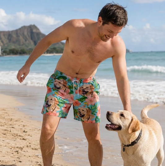 Man op het strand in een kleurrijke zwembroek met honden- en bloemenmotief. Labrador speelt naast hem in het zand.
