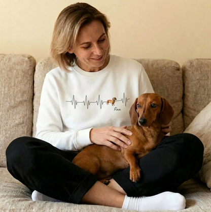 Witte sweater met hartslagmotief en een hond, lachende vrouw zit met een teckel op een bank.