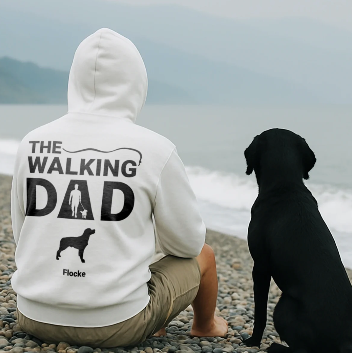 Witte hoodie met rugprint "THE WALKING DAD" en hondenmotief, gedragen door een man die op het strand zit.