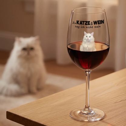 Gepersonaliseerd wijnglas met zwarte tekst en rode wijn, ernaast een witte kat op een houten tafel.