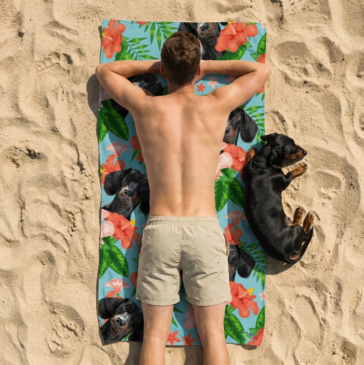 Persoon ligt op een gepersonaliseerde strandhanddoek met hondenmotieven, omringd door zand.