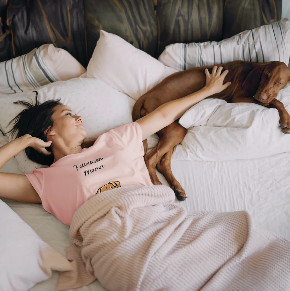 Vrouw in roze T-shirt ligt op bed naast een hond, omringd door kussens en een deken.