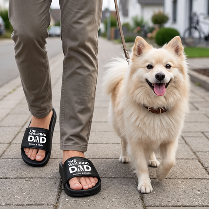 Persoon draagt gepersonaliseerde zwarte badslippers met opdruk "THE WALKING DAD" en loopt met een hond.